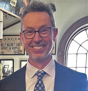 professor wearing a black suit, white shirt and blue tie in front of an oval window